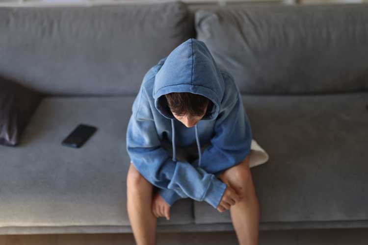 En tonårspojke sitter ensam på en grå soffa i ett vardagsrum och lutar sig framåt med böjt huvud, vilket förmedlar en stark känsla av stress, sorgsenhet och ensamhet