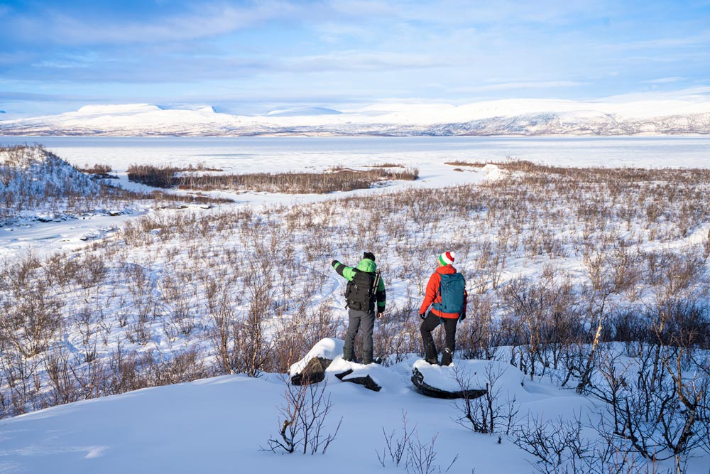 Två vandrare med ryggsäckar står på en snötäckt kulle och beundrar utsikten över en frusen sjö och ett vidsträckt arktiskt landskap i Abisko nationalpark